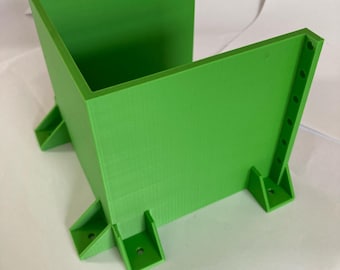 Puede incluir: Un contenedor verde brillante y angular con una abertura rectangular. El contenedor tiene cuatro patas de soporte y una serie de pequeños agujeros a lo largo de un lado. El objeto parece estar impreso en 3D.