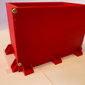 May include: A bright red rectangular planter box with brass-colored rivets and feet. The planter has a simple, modern design, suitable for indoor or outdoor use. The box is made of a solid material.