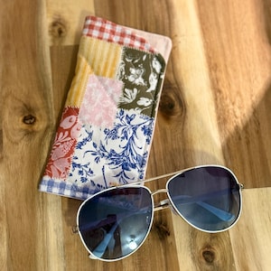 May include: A pair of aviator sunglasses with thin gold-tone metal frames and dark blue gradient lenses, positioned next to a rectangular quilted fabric case featuring a patchwork design of red, yellow, pink, green, and blue floral patterns.