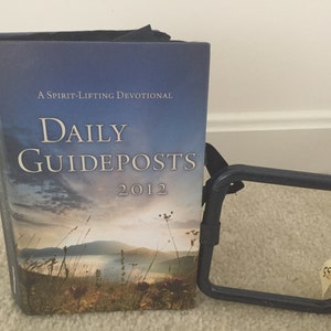 Peut inclure: Un livre à couverture rigide intitulé "Daily Guideposts 2012" avec une image de paysage sur la couverture. Le livre est à côté d'un cadre en métal bleu foncé avec une étiquette attachée. La couverture du livre présente un ciel bleu et un champ.