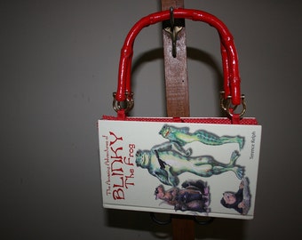 May include: A red book bag with a red handle. The book cover features a green frog with the title "The Amazing Adventures of Blinky the Frog" by Terence Ralph.
