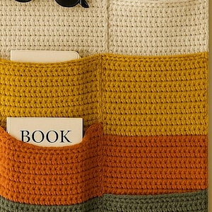 Op de afbeelding: Een crèmekleurige, gehaakte wandorganizer met het woord "kip" in zwarte letters. Het heeft zakken en is gestreept met geel, oranje en groen. Een zonnebril rust boven de zakken en een boek is zichtbaar.