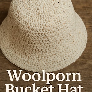 Op de afbeelding: Een crèmekleurige bucket hat gemaakt van een getextureerd, geweven materiaal. De hoed heeft een ronde kroon en een naar beneden hellende rand. De tekst "Woolporn Bucket Hat" staat in het wit onderaan de afbeelding.