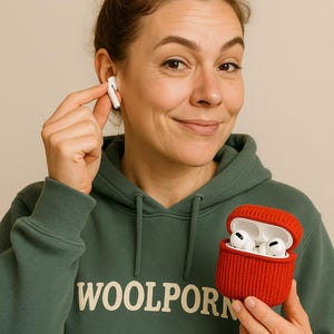 Op de afbeelding: Een vrouw in een groene hoodie met de tekst "WOOLPORN" houdt een rode gebreide hoes vast met witte draadloze oortelefoons. Eén oortelefoon zit in haar oor. De achtergrond is neutraal beige.