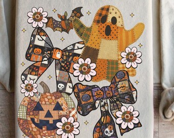 May include: Beige sweatshirt with a Halloween design. The design features a patchwork ghost, a jack-o'-lantern, and decorative bows with floral accents. A bat is also included in the design. The sweatshirt is folded.