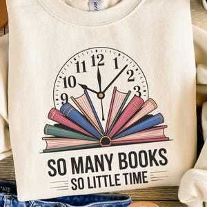 May include: Cream-coloured sweatshirt with a clock design featuring open books. The clock hands point to 12:01. The text "SO MANY BOOKS SO LITTLE TIME" is printed below the clock. The sweatshirt is folded on top of blue jeans and sandals.
