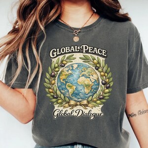 May include: Dark gray t-shirt with the words "Global Peace" and "Global Dialogue" above and below a globe illustration. The globe is surrounded by a wreath of olive branches and olives. The shirt is a casual style.