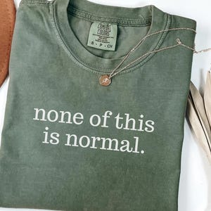 May include: An olive green t-shirt with the phrase "none of this is normal" printed in white. A gold-coloured necklace with a small pendant rests on the shirt. The shirt is folded on a white surface.