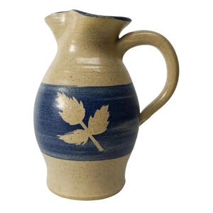 May include: A small, beige ceramic pitcher with a blue band and a white leaf design. The pitcher has a rounded body and a handle.