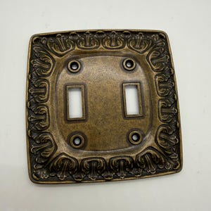 May include: A square double toggle light switch wall plate with an antique brass finish. The plate features a decorative border with a repeating leaf-like pattern and four screw holes positioned around two rectangular cutouts for switches.