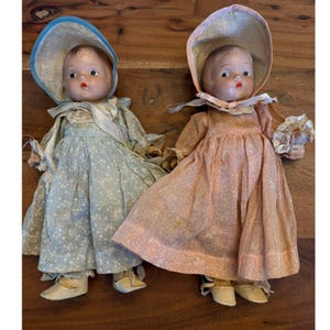 Peut inclure: Deux poupées anciennes avec des visages peints, vêtues de robes et de bonnets. L'une porte un bonnet bleu clair et une robe à motifs floraux. L'autre porte une robe et un bonnet roses. Les deux poupées ont des chaussures assorties.