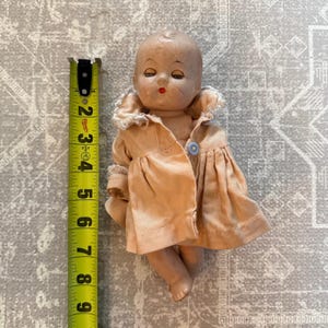 Puede incluir: Una muñeca vintage con un vestido color melocotón y un botón azul. La muñeca mide aproximadamente 10 cm de altura. La cara de la muñeca tiene rasgos pintados, incluyendo ojos cerrados y una boca roja. La muñeca es un artículo de colección.