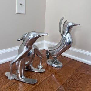 May include: Two polished silver penguin sculptures. One penguin is on skis, the other has its wings raised. The sculptures are on a hardwood floor, with a light-colored wall in the background. The penguin sculptures are decorative.