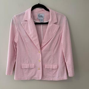 May include: Pink and white striped blazer with a notched collar, two front pockets, and three buttons. The blazer is hanging on a black hanger against a neutral wall. The label reads "Lilly Pulitzer".