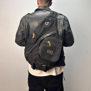 May include: Black crossbody bag with multiple zippered compartments and a gold-toned zipper. The bag is worn over the shoulder and across the chest. The bag has a small gold-toned logo on the front.