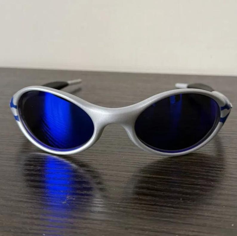Puede incluir: Gafas de sol plateadas con lentes azules. Las monturas ovaladas tienen un acabado plateado con detalles azules en las patillas. Las lentes son de un azul vibrante, ofreciendo un aspecto deportivo.