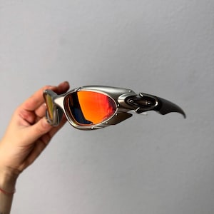 May include: Silver-framed sunglasses with vibrant red-orange lenses. The sporty eyewear features a sleek, aerodynamic design with a curved temple and a metallic finish. The lenses have a mirrored effect.