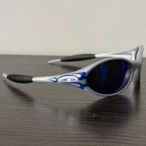 Puede incluir: Gafas de sol plateadas con lentes azules. Las monturas presentan un dise&ntilde;o de llamas en azul y blanco. Las patillas tienen puntas negras y un cuerpo plateado. Las lentes son de forma ovalada y azul oscuro.