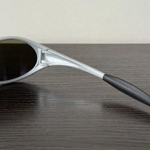 Puede incluir: Gafas de sol plateadas con lentes oscuras. Las gafas de sol tienen un dise&ntilde;o elegante y moderno con una montura curvada y una patilla negra. Las lentes son de forma ovalada y parecen tintadas.