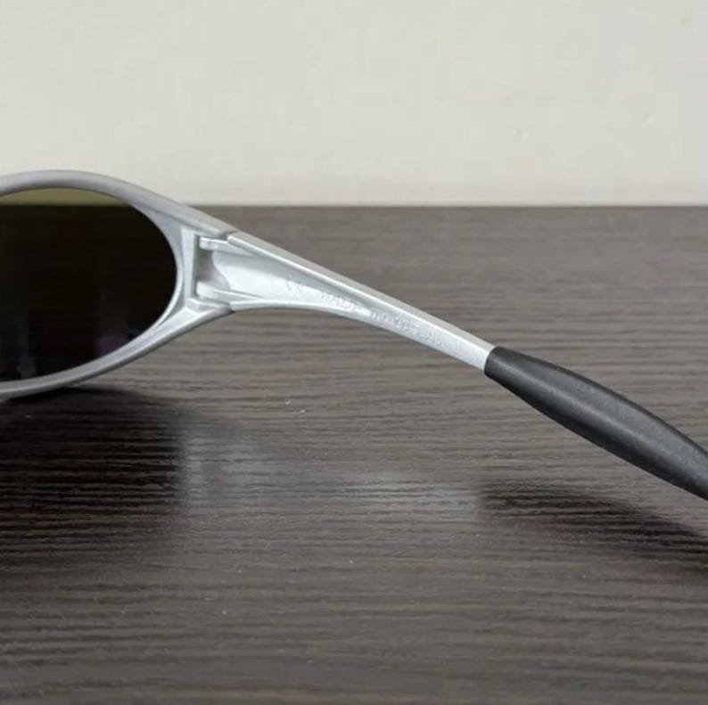 Puede incluir: Gafas de sol plateadas con lentes oscuras. Las gafas de sol tienen un dise&ntilde;o elegante y moderno con una montura curvada y una patilla negra. Las lentes son de forma ovalada y parecen tintadas.