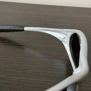 Puede incluir: Primer plano de unas gafas de sol con montura plateada y patillas negras. Las gafas de sol tienen un dise&ntilde;o moderno, con una montura curvada y lentes oscuras. El nombre de la marca es visible en la montura.