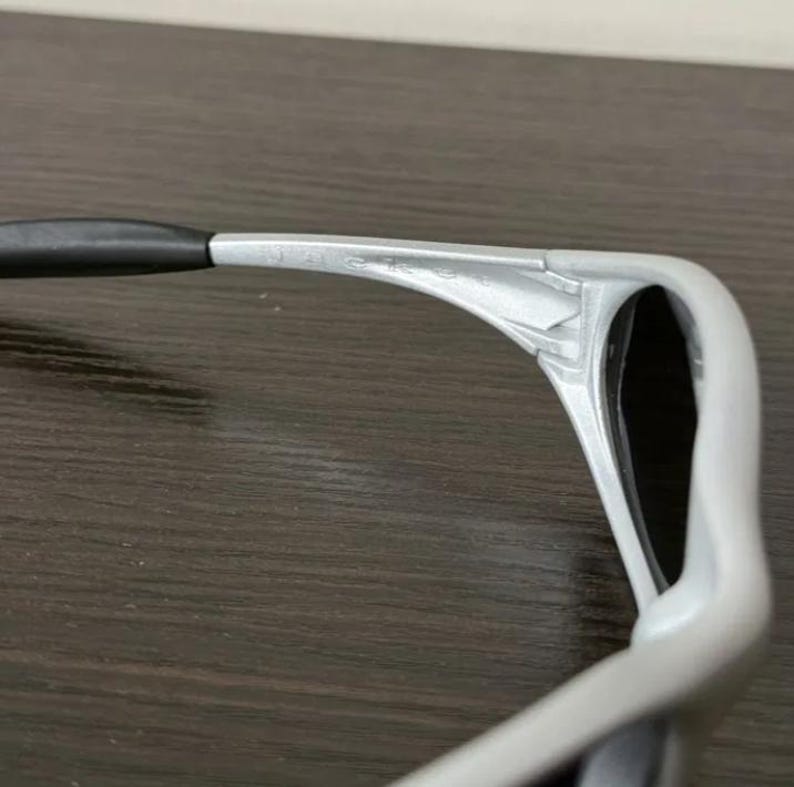 Puede incluir: Primer plano de unas gafas de sol con montura plateada y patillas negras. Las gafas de sol tienen un dise&ntilde;o moderno, con una montura curvada y lentes oscuras. El nombre de la marca es visible en la montura.