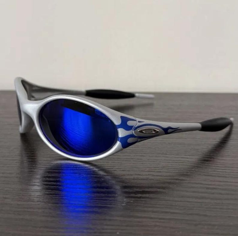 Puede incluir: Gafas de sol plateadas con lentes azules. Las patillas presentan un dise&ntilde;o de llama azul. Las gafas tienen patillas negras y un dise&ntilde;o elegante y moderno. Las lentes reflejan el entorno.