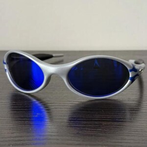 Puede incluir: Gafas de sol plateadas con lentes azul oscuro. Las monturas tienen un dise&ntilde;o curvo y detalles azules en forma de llama en las patillas. Las gafas de sol est&aacute;n sobre un fondo neutro.