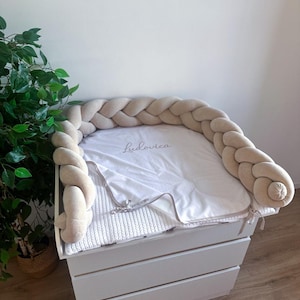 Op de afbeelding: Een witte commode met drie lades, bekroond met een wit gebreid kussen en een zachte, gevlochten bumper in een neutrale beige kleur. Een witte deken met de naam "Ludovica" ligt erop.