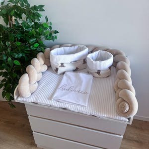 Può includere: Un fasciatoio bianco con tre cassetti, sormontato da un paraurti intrecciato color crema e un fasciatoio abbinato. Due cestini bianchi con un motivo a orsetti sono appoggiati sul fasciatoio, insieme a un panno bianco ricamato con il nome "Ludovica".