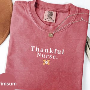 Może przedstawiać: Karmazynowa koszulka z napisem "Thankful Nurse." i grafiką plastra. Koszulka ma okrągły dekolt i krótkie rękawy. Srebrny naszyjnik z wisiorkiem jest zarzucony na koszulkę. Brązowy kapelusz jest widoczny w lewym górnym rogu.