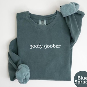 Può includere: Una felpa girocollo blu abete con la scritta "goofy goober" in lettere bianche. Le maniche lunghe sono arrotolate. La felpa è ripiegata ordinatamente su una superficie bianca.