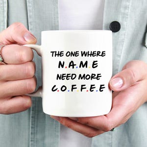 Könnte beinhalten: Weiße Keramik-Kaffeetasse mit schwarzem Text, der lautet: "THE ONE WHERE NAME NEED MORE COFFEE". Das Wort "NAME" ist schwarz, und die Buchstaben sind blau, gelb und rot gefärbt. Die Tasse wird von einer Person gehalten, die ein hellgrünes Hemd trägt.