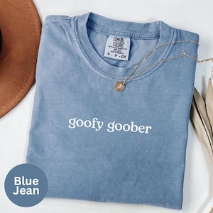 Può includere: Una maglietta blu jeans con le parole "goofy goober" stampate in bianco. La maglietta ha un girocollo e maniche corte. Una collana d'oro con un ciondolo è drappeggiata sulla maglietta.