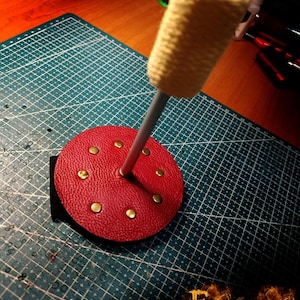 Puede incluir: Un accesorio de fuego de cuero rojo con remaches de latón, una correa negra y un mango amarillo. El accesorio está sobre una esterilla de corte verde con un patrón de cuadrícula. El logotipo "Furious Fire Lab" es visible.