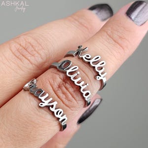 May include: Three silver rings with the names "Grayson", "Olivia", and "Kelly" engraved on them. The rings are stacked on a finger.