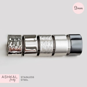 Peut inclure: Cinq bagues en acier inoxydable, chacune avec une texture différente. Les bagues mesurent 9 mm de large et sont disposées en rangée de gauche à droite. Les bagues sont toutes de couleur argent. Le texte "ASHKAL Jewelry" et "STAINLESS STEEL" est visible en bas de l'image.