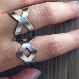 May include: Two silver-colored rings with an X and diamond-shaped cutout design. The rings are worn on fingers, showcasing a modern, geometric style. The rings are made of a shiny metal.