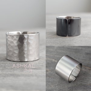 May include: Three silver rings, one with a hammered finish, one with a smooth finish, and one with a black oxidized finish. The rings are all wide and flat, and they are made of metal. The rings are on a gray wooden surface. ASHKAL Jewelry.