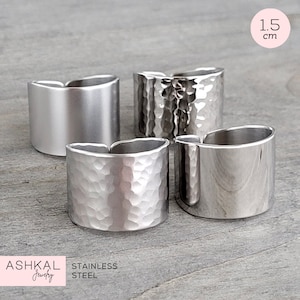 May include: Four silver rings, two with a hammered texture and two with a smooth finish. The rings are 1.5 cm wide. The rings are made of stainless steel. The text "ASHKAL Jewelry" and "STAINLESS STEEL" is visible in the image.