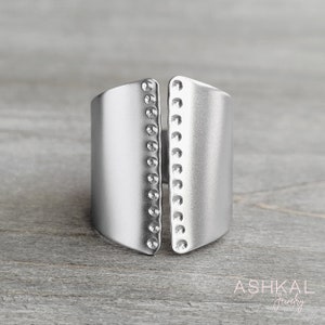 May include: A silver ring with a wide band and a split design. The band has a row of small circles on each side.