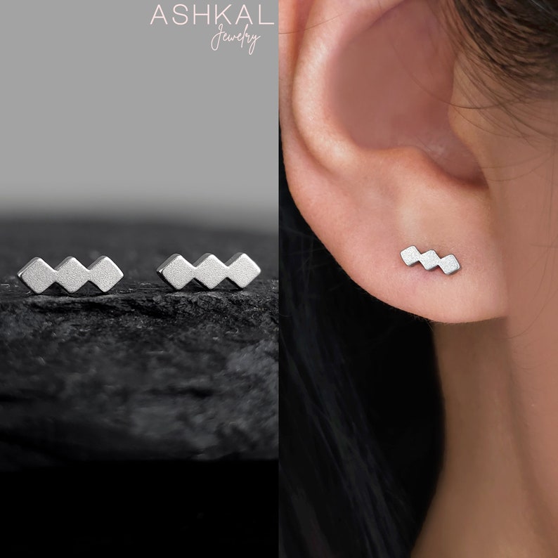 May include: A pair of silver geometric stud earrings. The earrings are shaped like a zig-zag pattern. The earrings are shown on a model's ear.