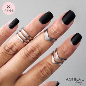 Peut inclure: Trois bagues argentées sur une main avec un vernis à ongles noir. Les bagues sont un ensemble de trois bandes fines et deux bagues en forme de chevron.