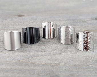 May include: Five silver and black wide band rings on a wooden surface. The rings are all different styles, including smooth, hammered, and textured.