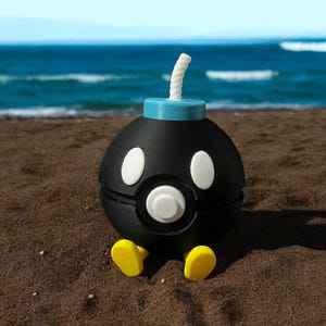 Puede incluir: Un personaje de bomba de dibujos animados negro con ojos blancos y una mecha blanca, sobre una escena de playa con un océano y un cielo azules. La bomba tiene pies amarillos y una parte superior azul claro.