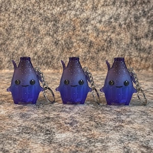 May include: Three identical translucent purple keychains shaped like small creatures with a gradient effect, glittery accents, two black circular eyes, and a simple smile. Each keychain features a metal chain and a circular split ring for attachment.