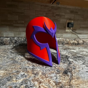 Könnte beinhalten: Ein roter und violetter Helm, der an einen Comic-Charakter erinnert. Der Helm hat eine abgerundete Oberseite mit einem violetten, stilisierten Design auf der Vorderseite und den Seiten. Hergestellt aus hartem Material.