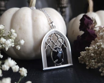 May include: A silver pendant necklace featuring a gothic arch design with an oval, faceted black gemstone. The pendant hangs on a delicate silver chain. The background includes white pumpkins and dried flowers, creating a gothic aesthetic.