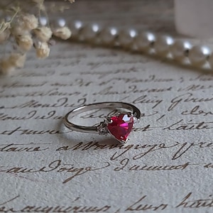 May include: A silver ring with a heart-shaped ruby gemstone and two smaller clear gemstones on the band.