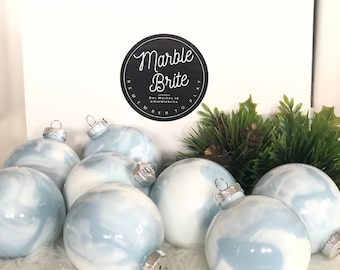 Puede incluir: Un conjunto de adornos navideños con efecto mármol en tonos azul claro y blanco. Cada adorno tiene una tapa plateada. Los adornos se exhiben sobre una superficie blanca y esponjosa, con una caja en el fondo con el texto "Marble Brite".